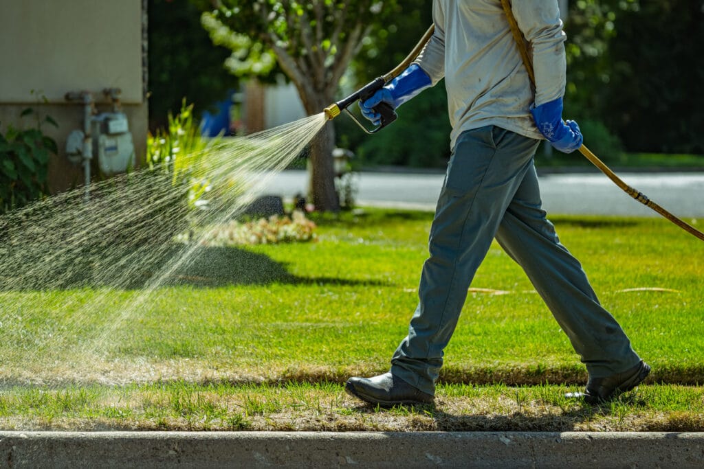 Technician applies weed control and fertilizer service with hose sprayer along curb edge of a Davis County Utah lawn.