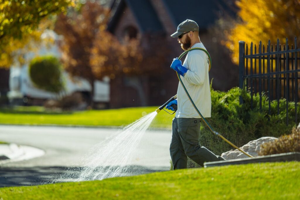 A lawn care technician sprays liquid treatment across a lush green lawn in a residential neighborhood.