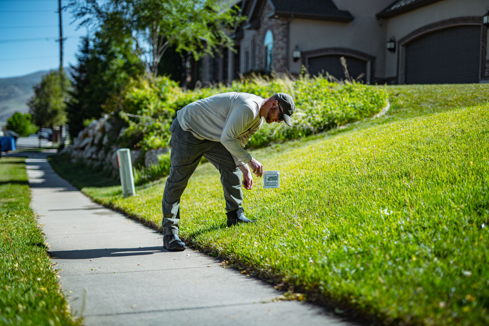 A lawn care technician bending down to place a small sign into a neatly maintained sloped green lawn beside a sidewalk.