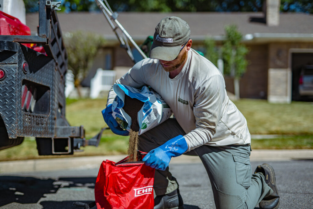 A lawn care technician kneeling beside a truck, pouring granular material from a bag into a red spreader hopper.