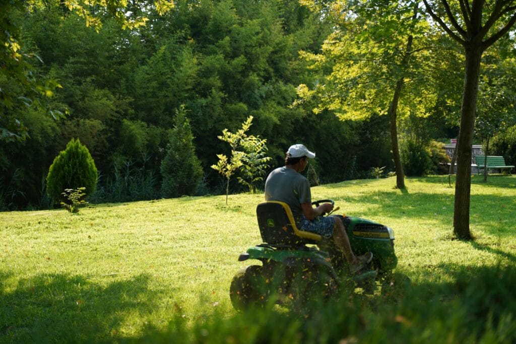 Person riding a yellow and green lawn mower cutting grass in a sunny residential yard